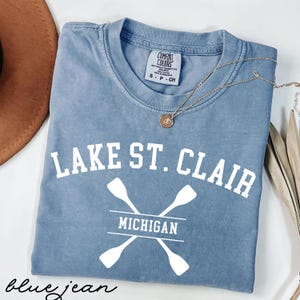 May include: Blue jean colored t-shirt with the text "LAKE ST. CLAIR MICHIGAN" in white. The design includes crossed oars. A gold necklace with a pendant is draped across the shirt. The shirt is a Comfort Colors brand.
