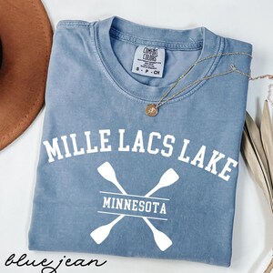 以下が含まれることがあります： 「MILLE LACS LAKE」の白い文字が入った、ブルーデニムカラーのTシャツ。その下に、2つの白いパドルが交差したデザインと「MINNESOTA」の文字。金のネックレスがシャツの上にかけられています。