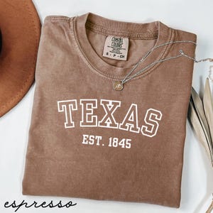 May include: Espresso-colored t-shirt with the word "TEXAS" in white block letters and "EST. 1845" underneath. A silver necklace with a pendant is draped across the shirt. The shirt is a Comfort Colors brand.