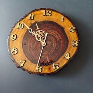 May include: A round wall clock made from a natural wood slice with a dark brown centre and a lighter brown outer ring. The clock features gold-coloured numbers from 1 to 12 and ornate gold-coloured metal clock hands set against the wood grain.