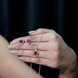 Puede incluir: Un conjunto de joyería compuesto por un collar y dos pendientes. Cada pieza presenta una piedra roja en forma de corazón rodeada por un halo de pequeñas piedras blancas engastadas en metal plateado. El colgante incluye un diseño de infinito.