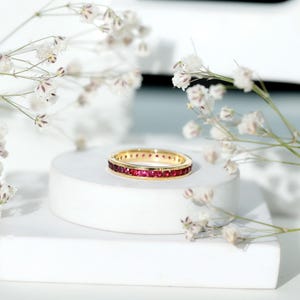 Könnte beinhalten: Ein goldener Bandring mit einer Reihe roter Edelsteine. Der Ring wird auf einem weißen Sockel mit weißen Blumen im Hintergrund präsentiert. Der Ring ist im Full-Eternity-Stil gehalten, wobei die Edelsteine das gesamte Band umschließen.