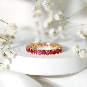 Könnte beinhalten: Ein goldener Ring mit einem durchgehenden Band aus leuchtend roten Edelsteinen. Der Ring liegt auf einer weißen Oberfläche, mit zarten weißen Blüten im Hintergrund. Das Design des Rings ist schlicht und elegant.