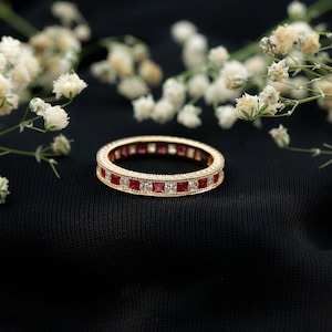 Op de afbeelding: Een goudkleurige eternity ring met afwisselend rode en heldere vierkante stenen. De ring is geplaatst tegen een zwarte achtergrond, met witte bloemen op de achtergrond.