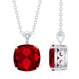 Peut inclure: Un collier en argent avec un grand pendentif en pierre précieuse rouge foncé taillée en coussin. Le pendentif est serti dans un cadre argenté avec des filigranes décoratifs sur les côtés. La chaîne est une fine chaîne en argent.