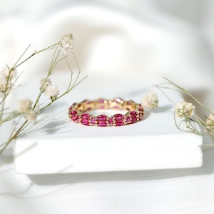 May include: A gold ring with a continuous band of oval-cut, vibrant pink gemstones. The ring is set on a white surface, with delicate white flowers and stems in the background, creating a soft, elegant aesthetic.