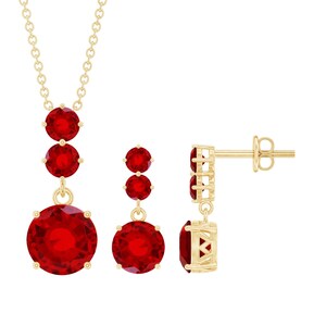 Könnte beinhalten: Ein goldfarbenes Schmuckset mit Halskette und Ohrringen. Die Halskette hat eine Kette mit drei roten Edelsteinen, der größte unten. Die Ohrringe haben zwei rote Edelsteine. Das Schmuckset ist vor weißem Hintergrund.