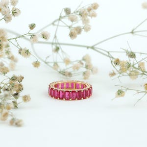 Könnte beinhalten: Ein goldfarbener Ring mit einem Band aus rechteckigen, leuchtend rosa Edelsteinen. Der Ring ist vor einem weißen Hintergrund mit zarten, weißen floralen Akzenten platziert, was einen reizvollen Kontrast erzeugt. Das Design des Rings ist elegant und auffällig.