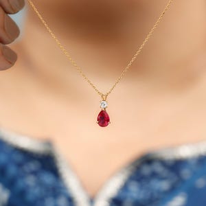 Puede incluir: Un collar de oro con un colgante con una piedra preciosa roja oscura en forma de pera. Encima del rubí hay una pequeña piedra preciosa redonda y transparente. El collar está en una fina cadena de oro. El fondo es una tela azul estampada.