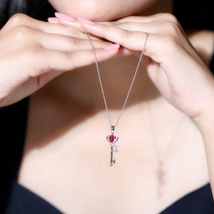 May include: A silver necklace with a key pendant. The key pendant has a red heart-shaped gemstone at the top, with a small diamond and a key design. The necklace is shown against a dark background.