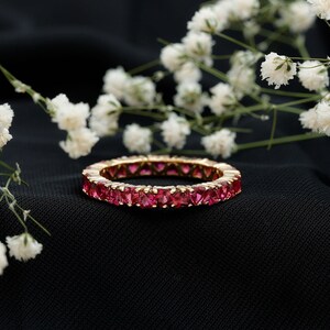 May include: A gold eternity ring featuring a continuous row of bright pink gemstones. The ring is presented on a black backdrop, with white flowers subtly placed in the background. The gemstones are square-cut and closely set around the band.