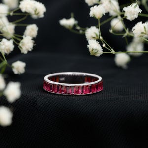 Op de afbeelding: Een zilveren ring met een band van rechthoekige, helder roze edelstenen. De ring staat tegen een zwarte achtergrond, met witte bloemen op de achtergrond. Het is een sieraad.