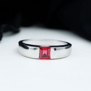 May include: A silver ring with a rectangular, bright red gemstone set in the center. The ring has a smooth, polished finish and a simple, elegant design. The gemstone is a vibrant, eye-catching color.