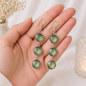 Pode incluir: Um par de brincos pendentes com três pedras redondas facetadas em verde claro, engastadas em metal prateado. Cada brinco pende de um gancho prateado simples e consiste em três engastes circulares alinhados verticalmente com gemas verdes.
