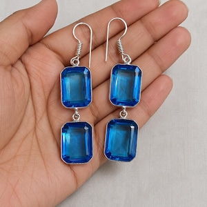 Pode incluir: Brincos pendurados prateados com duas pedras preciosas retangulares azuis vibrantes. As pedras são conectadas por um pequeno elo prateado e suspensas de um gancho prateado com um design em espiral. Os brincos são exibidos em um fundo neutro.