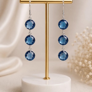 Pode incluir: Um par de brincos pendentes de tom prateado, cada um com três pedras circulares facetadas de cor azul-escura em engastes de metal. Os brincos pendem de ganchos prateados e são exibidos num suporte em forma de T dourado com uma base de mármore branco.