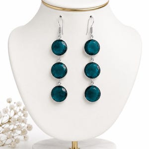 May include: A pair of dangle earrings featuring three circular, faceted, deep teal-colored stones set in silver-toned metal frames. Each earring hangs from a silver-toned hook, with the stones arranged vertically in a linear, drop-style design.