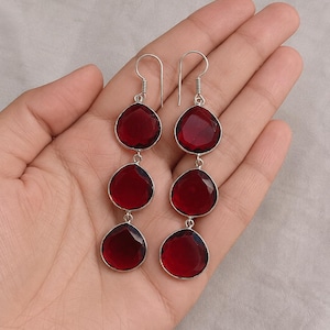 Pode incluir: Um par de brincos pendentes com três pedras vermelho-escuras facetadas em forma de gota, dispostas verticalmente numa armação de metal prateado. Cada brinco está suspenso por um gancho simples de metal prateado.