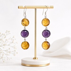 Pode incluir: Um par de brincos pendentes com três pedras preciosas redondas facetadas alinhadas verticalmente em engastes prateados. Cada brinco apresenta um citrino amarelo no topo, uma ametista roxa no centro e um citrino amarelo na parte inferior.