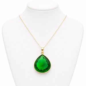 Op de afbeelding: Een goudkleurige ketting met een grote, druppelvormige, smaragdgroene hanger. De hanger is gezet in een gouden montuur en hangt aan een fijne gouden ketting. De ketting wordt getoond op een witte mannequin.