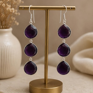 Pode incluir: Brincos pendurados com três pedras de ametista roxa escura facetadas em forma de gota. As pedras são engastadas em metal prateado e penduradas em ganchos de orelha prateados. Os brincos são exibidos em um suporte de joias dourado.