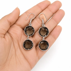 Pode incluir: Um par de brincos pendentes com duas pedras preciosas de quartzo fumê redondas e facetadas, engastadas em metal prateado. Cada brinco consiste em duas pedras circulares empilhadas verticalmente, suspensas por um gancho prateado simples.