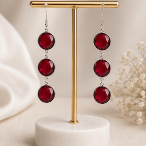 Pode incluir: Um par de brincos pendentes com três pedras circulares facetadas em vermelho profundo, engastadas em armações de metal prateado. Os brincos pendem de ganchos prateados num suporte dourado em forma de T com base de mármore branco.