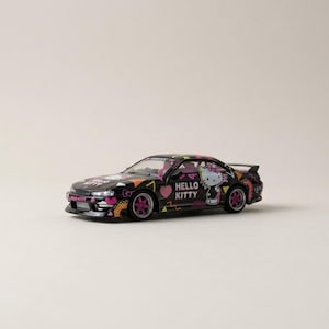 May include: A black toy car with Hello Kitty graphics. The car features pink and orange accents, with the Hello Kitty character prominently displayed on the side. The wheels are pink, and the car has a spoiler.