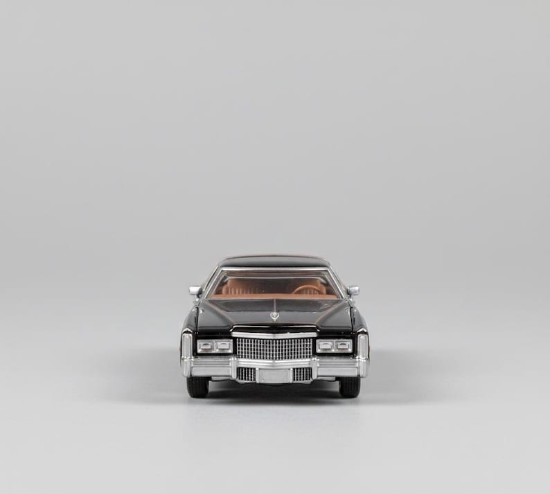 May include: A black classic car model with a tan interior. The front of the car features a chrome grill and headlights. The car is set against a plain gray background.
