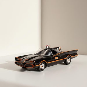 May include: A black toy car with orange trim, featuring a Bat symbol on the side. The car has a sleek design with a low profile, two clear windshields, and a red light on top. The wheels are black with silver rims.