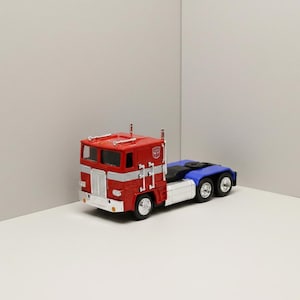 May include: A red and silver semi-truck toy with a blue trailer. The truck has a white grill and silver stripes. The cab features the Autobots logo. The toy truck is a collectible item.