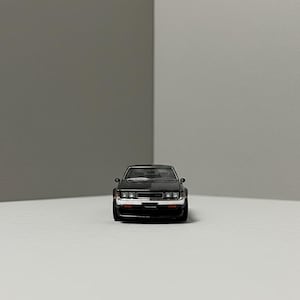 May include: A black diecast model car with a detailed front grille, headlights, and a prominent front bumper. The miniature vehicle is set against a neutral background, showcasing its sleek design and classic aesthetic. The car's body is a dark color, with silver accents.
