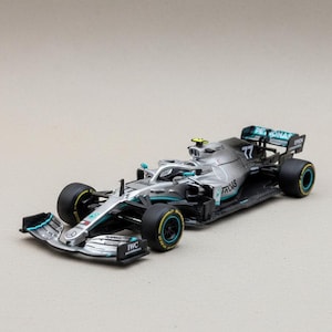 May include: A silver and black Formula 1 race car model with teal accents. The model features the number 77 and sponsor logos. The tires are black with yellow and teal details. The car is a detailed replica.
