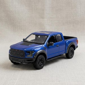 May include: A blue Ford Raptor pickup truck model with black wheels and trim. The model has a detailed front grille with the Ford logo and a black interior. The truck is a miniature replica of a full-size vehicle.