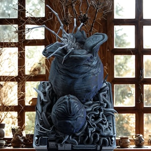Puede incluir: Una escultura azul oscuro con una forma bulbosa, detalles intrincados y una superficie texturizada. La escultura está adornada con elementos en forma de rama y se asienta sobre una base rectangular con rasgos enrollados en forma de tentáculos. El fondo incluye una ventana.