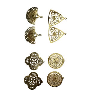 May include: An assortment of six ornate, filigree metal charms in gold and bronze tones. The charms include fan-shaped, triangular, clover-shaped, and circular designs, each with a small loop for attachment. These decorative elements are suitable for jewelry making or craft projects.