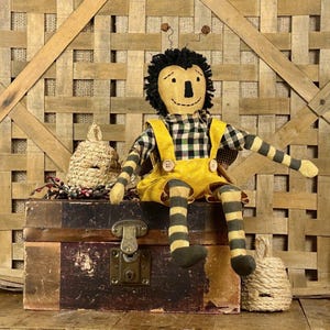 May include: A fabric bee doll with a black yarn head, yellow face, and black and yellow striped limbs. The doll wears a black and white checkered shirt under yellow overalls with two buttons. It sits on a weathered wooden trunk with a metal latch.
