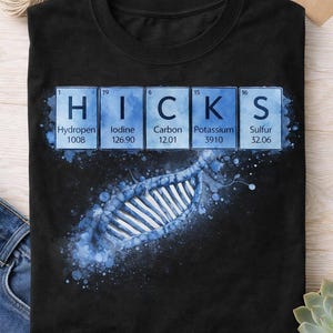May include: Black t-shirt featuring the word "HICKS" spelled out using periodic table elements: Hydrogen, Iodine, Carbon, Potassium, and Sulfur. A blue watercolor-style illustration of a spine is below the text.