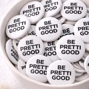 Puede incluir: Un conjunto de botones redondos blancos con el texto negro "BE PRETTI GOOD", presentados en un cuenco blanco. Los botones son brillantes y tienen un diseño sencillo y audaz. La imagen está bien iluminada y los botones son el foco principal.