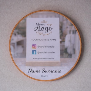 May include: A circular wooden framed mirror with a business card inside. The card displays a logo, business name, social media details, website, and space for a name and date. The background is a blurred room scene.
