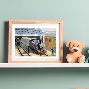 May include: A framed horizontal print featuring a blue steam locomotive with a face, positioned on train tracks inside a station. The illustration depicts a railway scene with a platform, a yellow carriage, and figures standing near the engine.