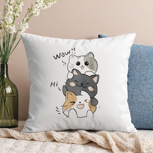 Könnte beinhalten: Weißes quadratisches Kissen mit einem Design von drei übereinander gestapelten Cartoon-Katzen. Die oberste Katze ist weiß und grau, die mittlere Katze ist grau und die untere Katze ist Schildpatt. Die Worte "Wow!!" und "Hi" sind auf dem Kissen geschrieben.