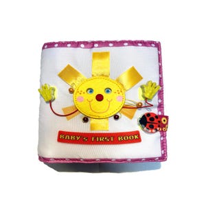 May include: A soft, white baby book with a pink polka dot border. The cover features a yellow sun with a smiling face, yellow hands, and the text "BABY'S FIRST BOOK" in red. A ladybug is attached to the side.