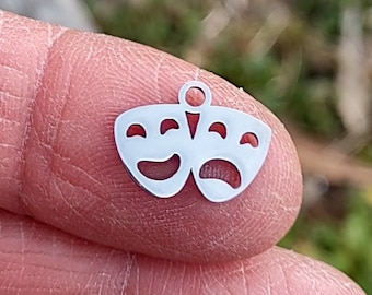 Puede incluir: Un charm de plata con forma de dos máscaras de teatro, una sonriente y otra frunciendo el ceño. El charm es pequeño y delicado, perfecto para añadir un toque de fantasía a un collar o una pulsera.