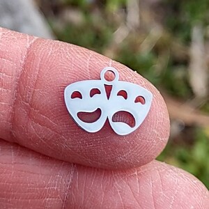 May include: A silver charm in the shape of two theater masks, one smiling and one frowning. The charm is small and delicate, perfect for adding a touch of whimsy to a necklace or bracelet.