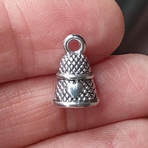 May include: A small, silver-colored thimble charm with a heart-shaped detail. The charm has a textured, diamond-patterned design on the upper and lower sections. It features a loop at the top for attaching to a necklace or bracelet. Ideal for jewelry making.