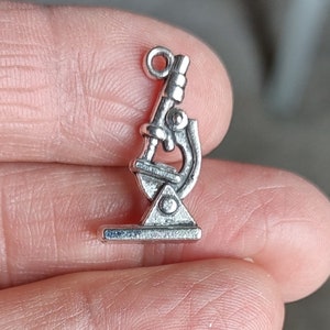 May include: A small, silver-colored microscope charm. The charm features detailed elements of a microscope, including the eyepiece, objective lenses, and base. The charm has a loop at the top for attaching to a necklace or bracelet. This charm is ideal for science enthusiasts.