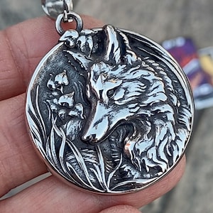 May include: A silver pendant featuring a detailed wolf head with a floral design. The wolf's head is facing the right side of the pendant and has a textured, realistic look. The floral design is on the left side of the pendant and features a cluster of leaves and flowers.