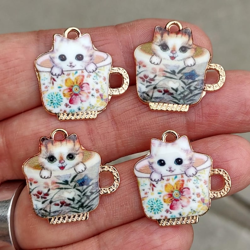 Puede incluir: Cuatro dijes con tem&aacute;tica de gatos. Cada dije presenta un gatito asom&aacute;ndose de una taza de t&eacute;. Las tazas de t&eacute; tienen patrones florales en varios colores, con bordes y asas doradas. Los dijes son probablemente para joyer&iacute;a o manualidades.