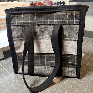 May include: A handmade tote bag with a patchwork design. The bag features a combination of gray plaid, herringbone, and solid fabrics. The bag has black trim and handles. The bag is approximately 12 inches tall, 14 inches wide, and 4 inches deep.